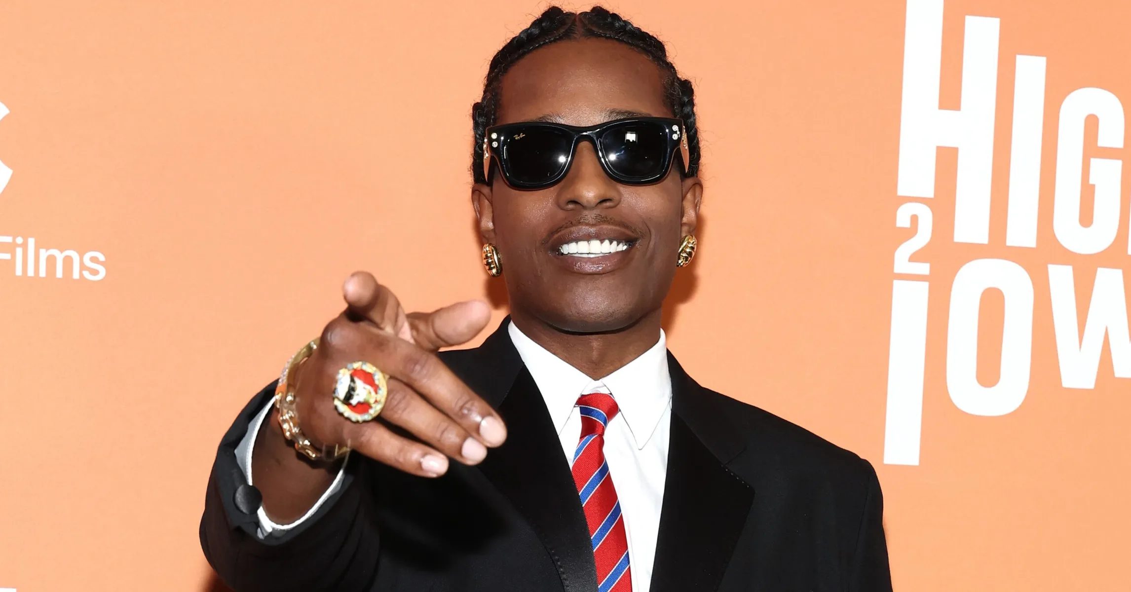 A$AP Rocky, wearing a black suit, white collared shirt, and a striped red and white tie, points toward the camera. He is wearing black sunglasses, large gold hoop earrings, and a prominent ring, against an orange and white promotional background.